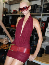 Woman wearing a red dress with a deep neckline and belt in a cluttered room.